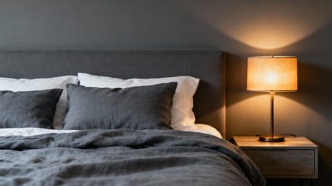 Camera da letto maschile minimalista con pareti grigio scuro, lenzuola in lino ardesia e una lampada con luce ambrata soffusa.